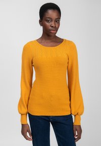 Frau in einem senfgelben Strickpullover mit gemustertem Yoke und Ballonärmeln, kombiniert mit dunkelblauen Jeans, vor einem hellen Hintergrund stehend.