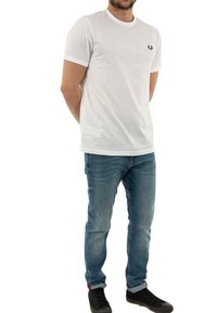 Camiseta blanca de manga corta con cuello redondo y logo negro en el lado izquierdo del pecho, combinada con jeans de mezclilla azul claros y zapatillas negras.