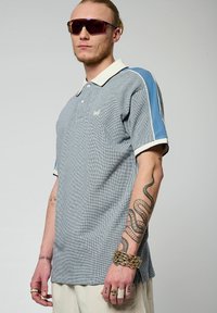 Polo shirt à motif à carreaux bleu et blanc, avec des manches bleues contrastantes et un col crème. Fabriqué en tissu texturé.