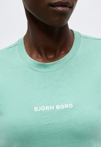 Κοντινό πλάνο ενός ατόμου που φοράει ένα ανοιχτοπράσινο t-shirt Björn Borg με λευκό λογότυπο στο κέντρο του στήθους.