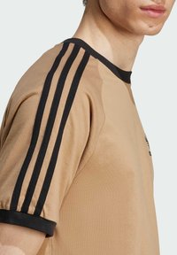 Camiseta de manga corta en tejido beige con cuello acanalado negro y tres rayas negras a lo largo de cada hombro, que presenta un pequeño logo.