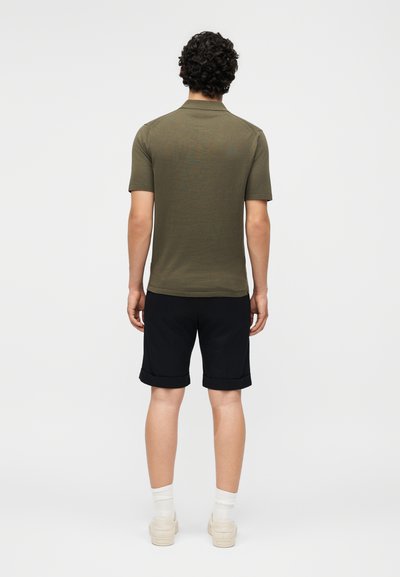 Άνδρας με σγουρά μαλλιά στέκεται με την πλάτη, φοράει πράσινο μπλουζάκι πόλο με κοντά μανίκια, μαύρα σορτς γυρισμένα, άσπρες κάλτσες και μπεζ αθλητικά παπούτσια με λευκό φόντο.