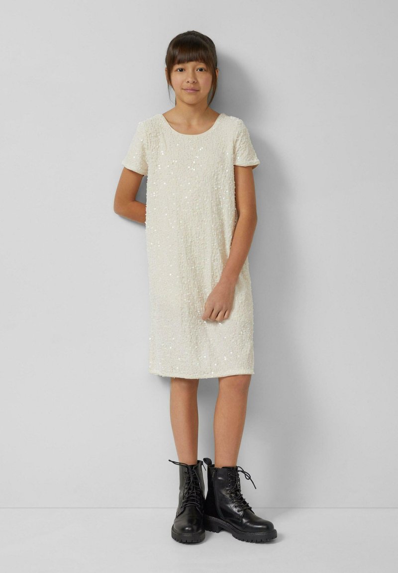 Kremowa sukienka z krótkim rękawem wykonana z teksturowanej tkaniny z cekinowymi akcentami, połączona z czarnymi sznurowanymi botkami za kostkę z grubymi podeszwami.