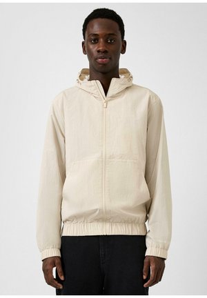 Junger Mann trägt eine hellbeige Kapuzenjacke mit Reißverschluss, elastischen Bündchen und Saum, steht vor einem schlichten weißen Hintergrund.