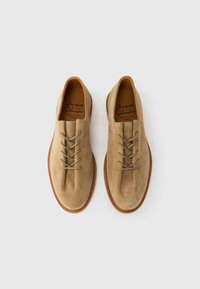 Chaussures en daim beige avec une texture lisse, bout rond et cinq paires d'œillets. Semelle extérieure en caoutchouc marron et étiquette de marque embossée visible à l'intérieur.