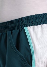 Cintura elástica en pantalones cortos deportivos verde azulado oscuro con forro de malla blanco y ribete verde azulado claro a lo largo de la costura lateral.