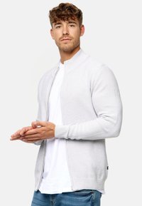 Cárdigan de cremallera gris claro con cuello y puños acanalados, que presenta un patrón de punto texturizado y bolsillos laterales, combinado con una camiseta blanca.
