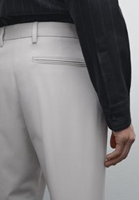 Helle graue Hosen haben eine glatte Textur und eine Gesäßtasche, die durch eine horizontale Naht betont wird, kombiniert mit einem dunkel gestreiften Hemd.