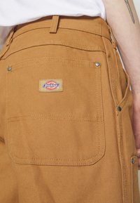 Brune arbejdsbukser lavet af struktureret stof med synlige søm, to baglommer og et Dickies-logo-patch nær taljen.
