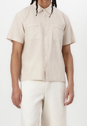 Mand iført en lysebeige kortærmet skjorte med to brystlommer og off-white bukser mod en ensfarvet baggrund.