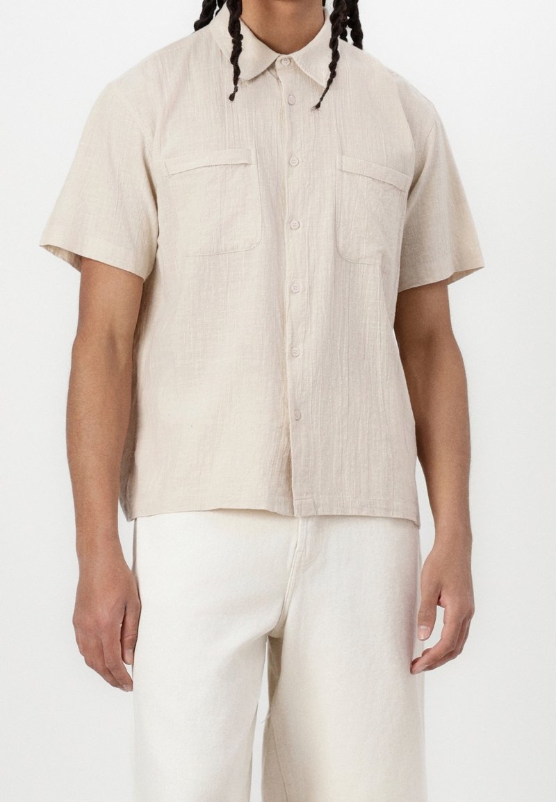 Homme portant une chemise beige clair à manches courtes avec deux poches poitrine et un pantalon blanc cassé sur un fond uni.