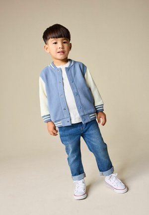 Ljusblå collegejacka med vita ärmar, tryckknappar, randiga muddar; vit t-shirt under; uppvikta jeans; vita sneakers.