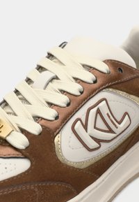 Καφέ σουεντ και δερμάτινο αθλητικό παπούτσι με εκρού κορδόνια, χρυσές λεπτομέρειες και λευκό λογότυπο. Διαθέτει ενισχυμένη μύτη και υφασμάτινη πλευρική επιφάνεια με υφή.