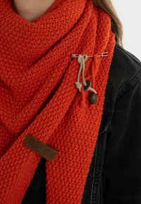 Gehaakte oranje sjaal met een textuurpatroon, afgewerkt met een decoratieve veiligheidsnaald en een tan label met "Knit Fraction" erop.