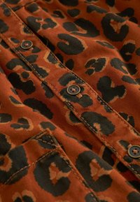 Tissu à imprimé léopard dans des tons chauds d'orange et de noir. Doté de boutons marron et d'une couture détaillée. Texture douce avec une poche visible.