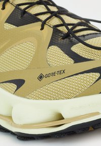 Zapatilla de correr GORE-TEX en malla de color amarillo claro con acentos negros. Presenta una parte superior texturizada, refuerzo en el talón y suela de amortiguación flexible.
