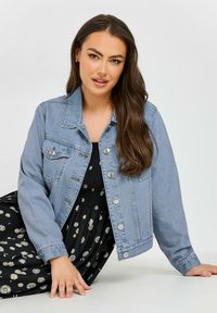 Giacca di jeans azzurro chiaro con chiusura a bottoni, due tasche sul petto e maniche leggermente sovradimensionate. Indossata sopra un vestito nero a fiori.