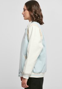 Svetlomodrá a krémová varsity bunda s rebrovanými manžetami, bočnými vreckami a vyšitým emblémom na hrudi. Mäkko tkanina a voľný strih.