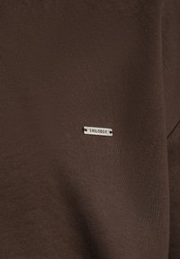 Liten silverfärgad rektangulär metalltag med "SMILODOX" präglat på mörkbrun ribbad textil.