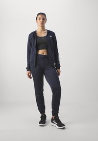 Námořnická sportovní souprava obsahující zipovou mikinu s logem, přiléhavý crop top a taperované tepláky, v kombinaci s černými sportovními botami.