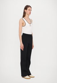 White tank top with dark trim featuring "MM" logo, paired with high-waisted black trousers and light-colored shoes.