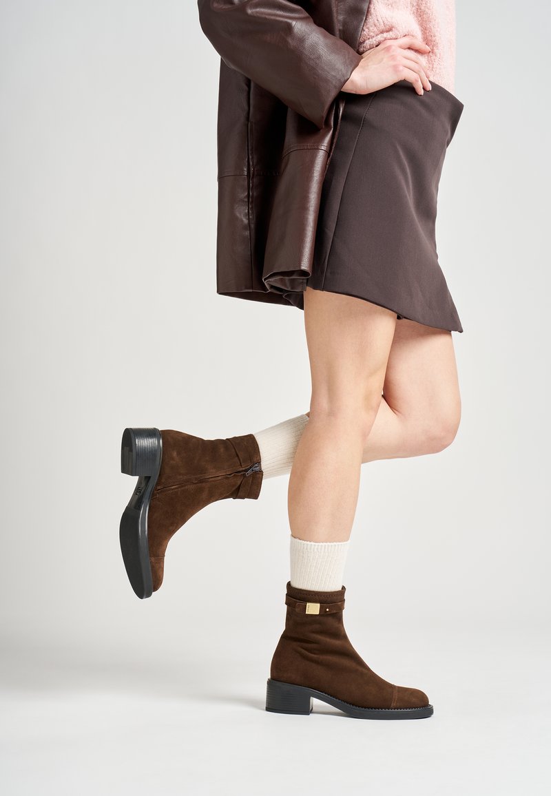 Stivaletti alla caviglia in suede marrone con tacco a blocco, dotati di una chiusura con zip e un accento metallico nella parte superiore, abbinati a calzini color crema chiaro.