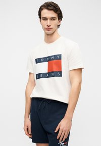 Κρεμ βαμβακερό t-shirt με σχεδιασμό λογότυπου σε ορθογώνιο, σε μπλε ναυτικό και κόκκινες αποχρώσεις, με το κείμενο "TOMMY JEANS". Φορεμένο με σορτς μπλε ναυτικού με κορδόνι.