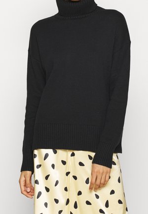 Pull à col roulé côtelé noir associé à une jupe en satin crème avec des taches noires irrégulières, porté par une personne sur fond blanc.