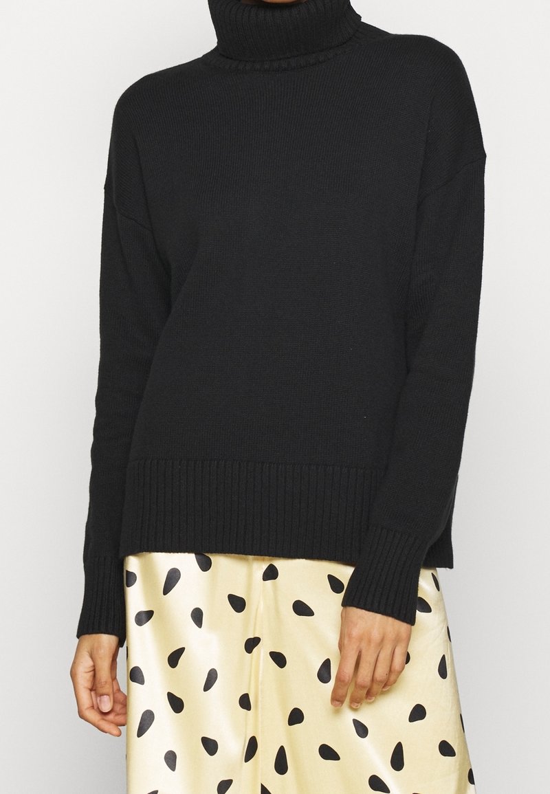 Pull à col roulé côtelé noir associé à une jupe en satin crème avec des taches noires irrégulières, porté par une personne sur fond blanc.