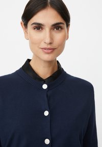 Marineblauwe cardigan met een ribgebreide halslijn, voorzien van vijf zilveren knopen, gedragen over een zwart overhemd met kraag. Een gladde textuur.