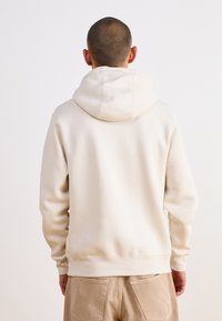 Beige hoodie med en mjuk textur, som har en avslappnad passform och ribbade ärmslut. Visad från baksidan, som visar detaljer kring halsringningen.