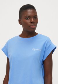 Lichtblauwe katoenen t-shirt met korte opgerolde mouwen. Kenmerkt zich door 'Day dreamer' geborduurd in het wit op de voorkant. Gladde textuur, ontspannen pasvorm.