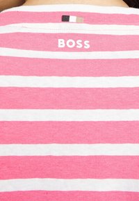 Růžová a bílá pruhovaná košile s nápisem "BOSS" vytištěným bílou barvou poblíž límce a malou černobílou a béžovou etiketou nad ní.