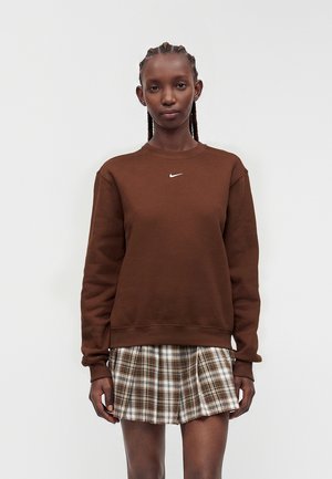 Vrouw met gevlochten haar die een bruine Nike-sweatshirt en een bruine geruite geplooide rok draagt, staand tegen een witte achtergrond.