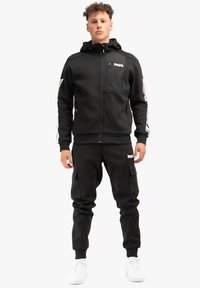 Melnā rāvējslēdzēja virsjakā ar priekšējo kabatu un baltām sānu svītrām, kopā ar melnām krāsu biksēm, kas ir ar sānu kabatām, un baltām kedām.