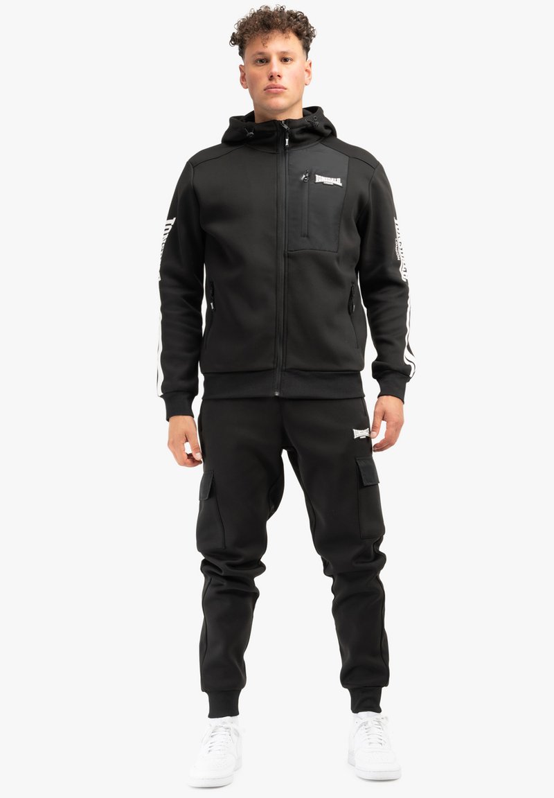 Melnā rāvējslēdzēja virsjakā ar priekšējo kabatu un baltām sānu svītrām, kopā ar melnām krāsu biksēm, kas ir ar sānu kabatām, un baltām kedām.