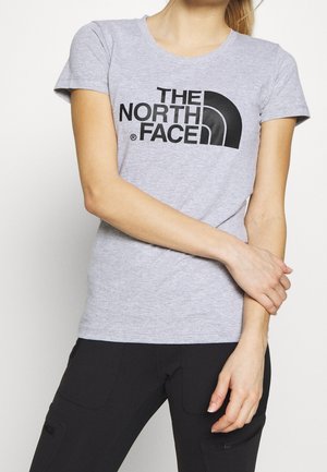 Femme portant un t-shirt gris clair à manches courtes avec le logo noir « THE NORTH FACE » et un pantalon noir, debout les bras croisés.