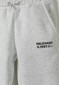 Grijze joggingbroek van zacht materiaal, voorzien van een elastische tailleband met trekkoord, pockets en zwarte tekst die zegt "RELEVANT S.1997 // --".