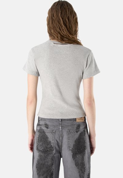 Femme aux cheveux bruns ondulés portant un t-shirt gris clair à manches courtes et un jean gris avec des taches foncées ressemblant à de la peinture sur les poches arrière et les jambes.