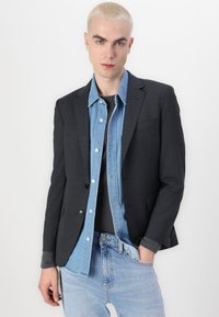 Temno sivi blazer z zareznimi reverji in enojnim gumbom, položen na svetlo modro denim srajco, v kombinaciji s svetlo modrimi kavbojkami.