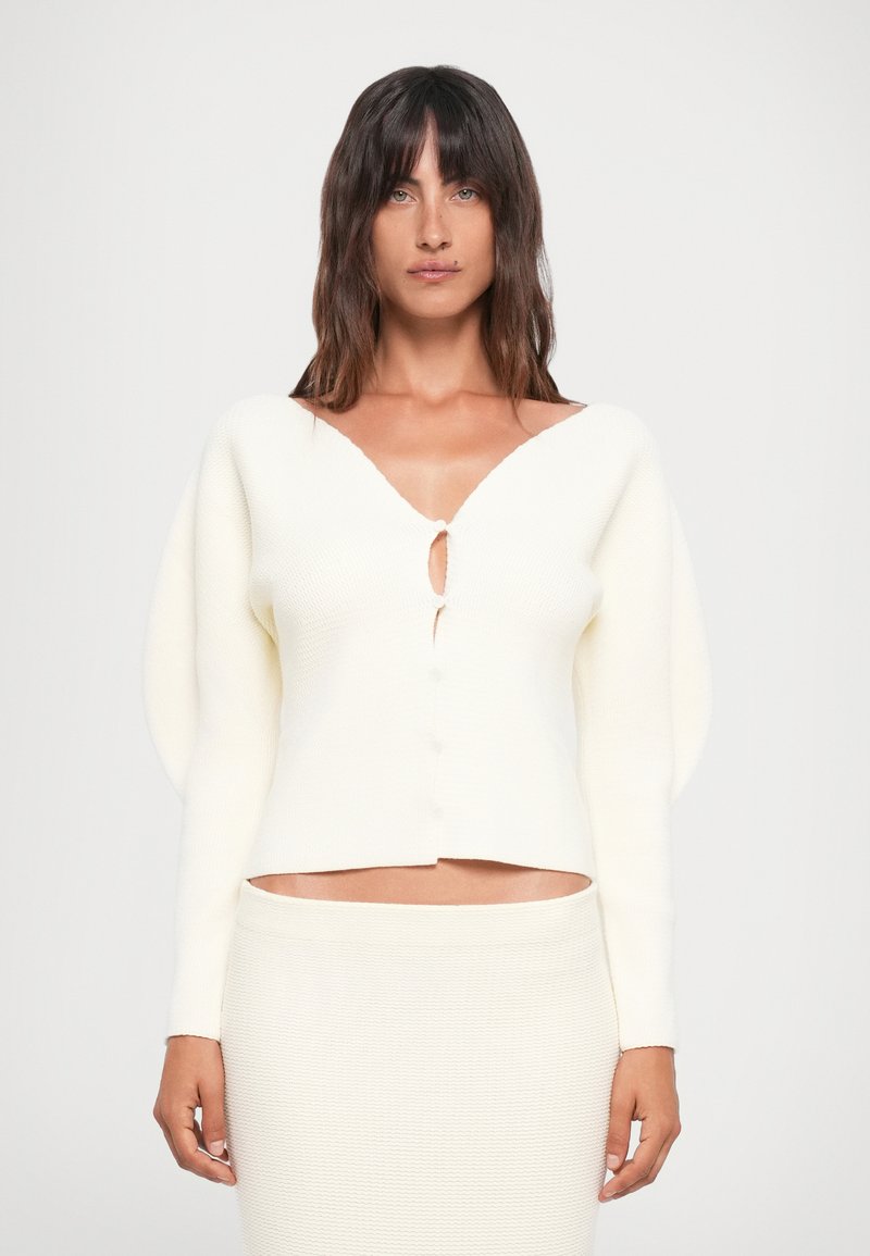 Bela pletena jakna z V-izrezom, spredaj z gumbi in napihnjenimi dolgimi rokavi. Tekstura je rebrasta, v kombinaciji z ujemajočo se krilom.