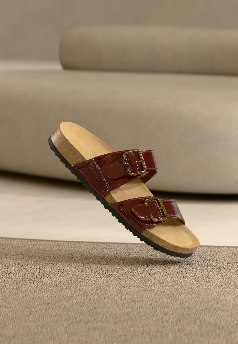 Sandale bordeaux à double bride avec boucles dorées, semelle en liège et semelle noire texturée sur fond de tapis beige et canapé.