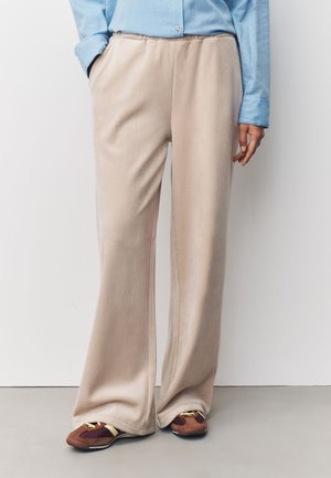 Person iført beige vidde kordbukserebukser, lyseblå skjorte med knapper og brune sneakers, der står mod en hvid væg.