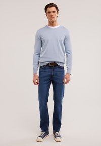 Suéter de cuello en V de un azul claro, con textura de punto medio, combinado con vaqueros de un azul oscuro y zapatillas azules con detalles en blanco. El modelo está de pie, mirando hacia adelante.