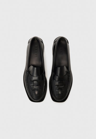 Mocassins en cuir noir avec un design tissé sur le bout, une forme arrondie et des détails de couture le long de la semelle.