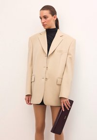 Béžový oversized blejzer so štruktúrovanými ramenami, dvoma prednými vreckami a tromi gombíkmi. Pod ním čierny top s vysokým golierom a bordová kabelka.