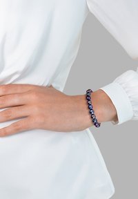 Bracciale di perle viola e blu con una finitura liscia e lucida, composto da perline rotonde infilate insieme. Indossato su un polso sopra una blusa bianca.
