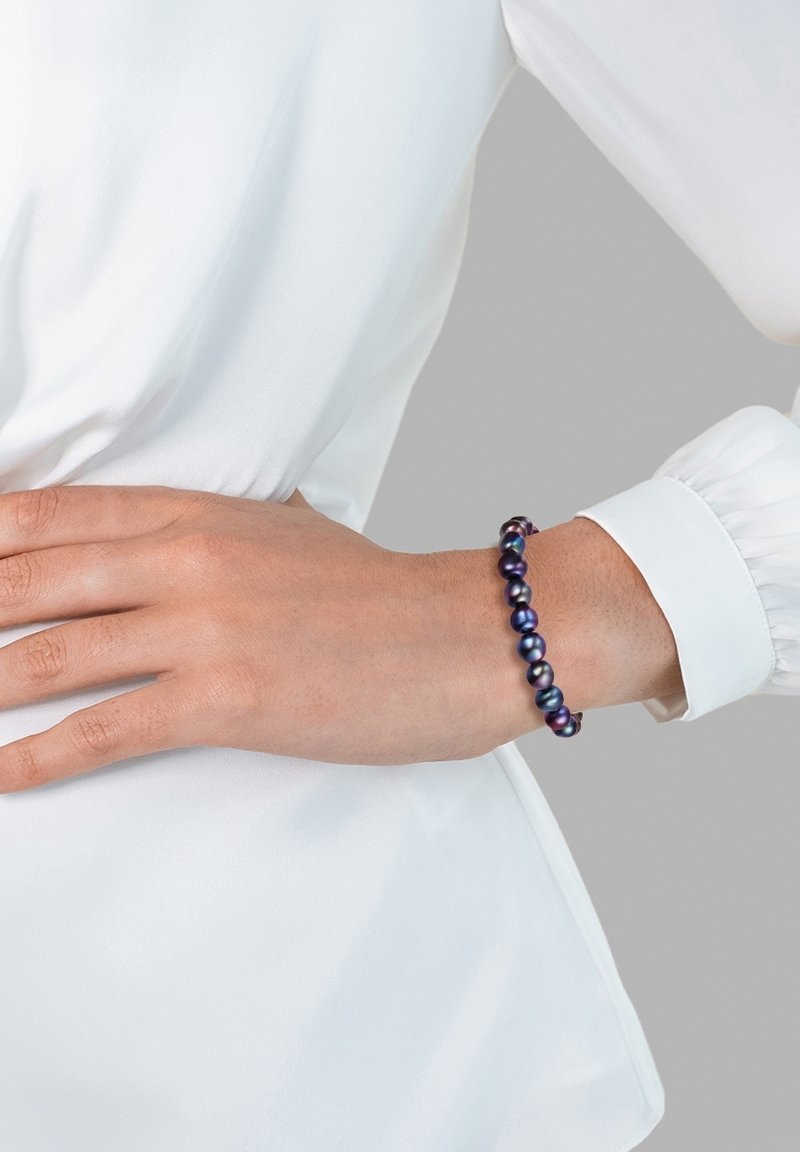 Bracciale di perle viola e blu con una finitura liscia e lucida, composto da perline rotonde infilate insieme. Indossato su un polso sopra una blusa bianca.