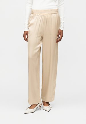 Beige, weit geschnittene Hosen mit elastischem Bund, kombiniert mit einem weißen Langarmshirt und weißen Slingback-Schuhen mit niedrigem Absatz und Schnallendetail.