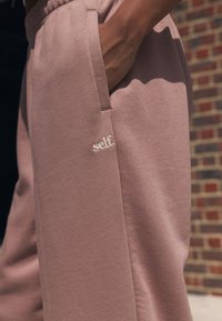 Lichtroze joggingbroek gemaakt van zachte stof, met zijzakken en een klein geborduurd "self." logo aan de voorkant.